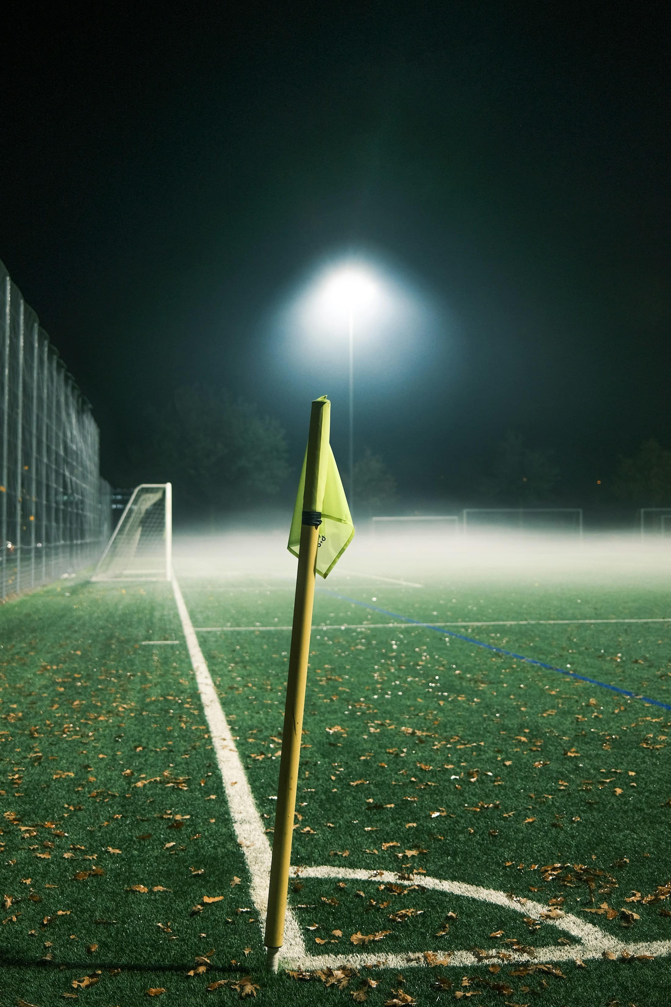 Eckfahne an einem Fußballplatz am Abend, im Hintergrund leuchtet das Flutlicht durch den Nebel