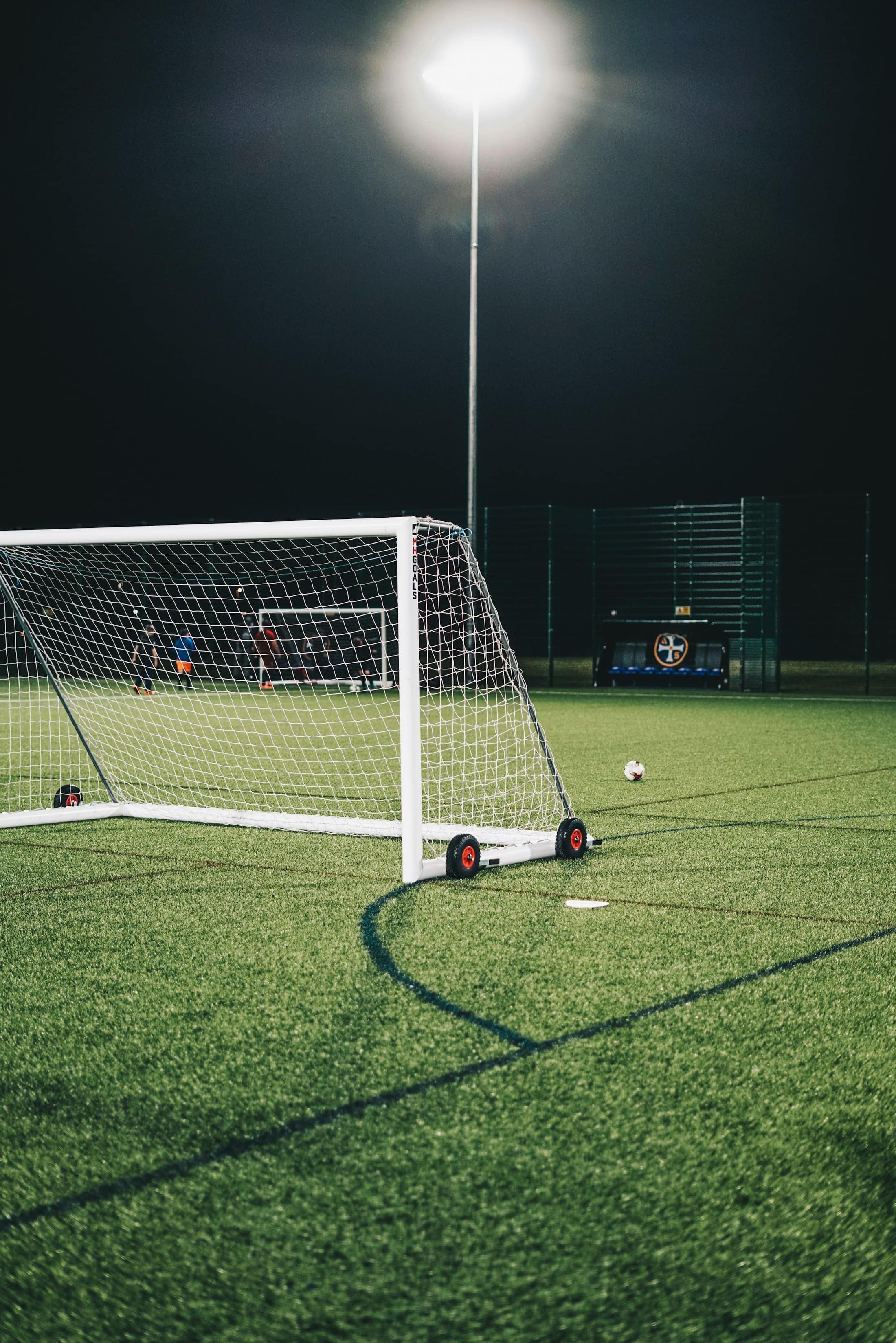 Ein Fußballtor auf einem Kunstrasenplatz, hell ausgeleuchtet von Flutlichtmasten in der Nacht